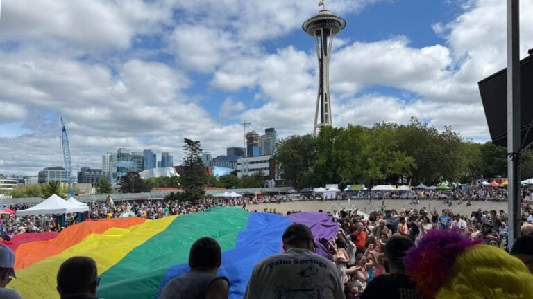 A large crowd unfurls a giant rainbow flag at Seattle PrideFest, with the Space Needle rising over Seattle Center under a partly cloudy sky.