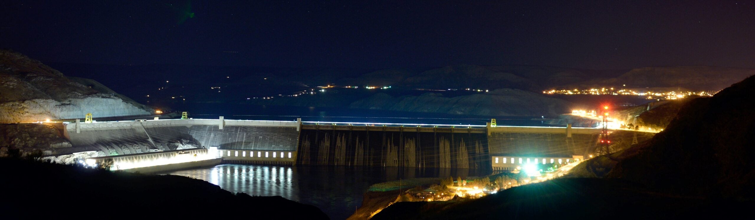 Grand Coulee Dam lit at night above the Columbia River, with Lake Roosevelt behind it and the lights of Grand Coulee to the right