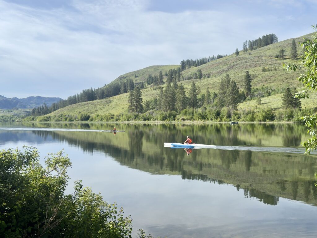 A kayaker and a rowing scull on the flat calm water of Pearrygin Lake, with the hills reflected on the surface