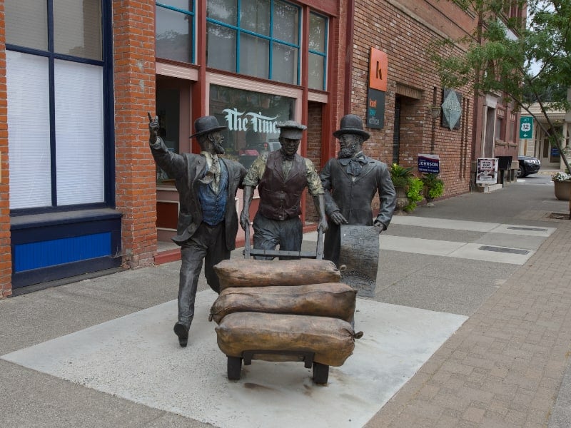 Three bronze figures pushing a hand cart loaded with sacks on a downtown sidewalk in Waitsburg, Washington, with brick storefronts behind them