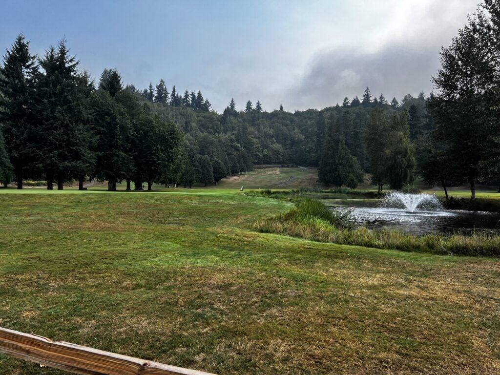 A wide fairway at Maplewood Golf Course with the pond fountain and the forested Cedar River valley hillside